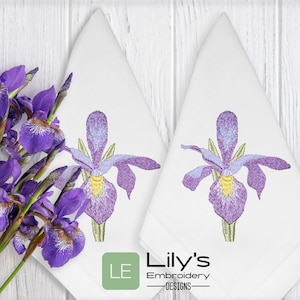 May include: Two white embroidered napkins with a purple iris flower design. The napkins are folded in a triangle shape and have a green label with the text "LE Lily's Embroidery -DESIGNS- ".