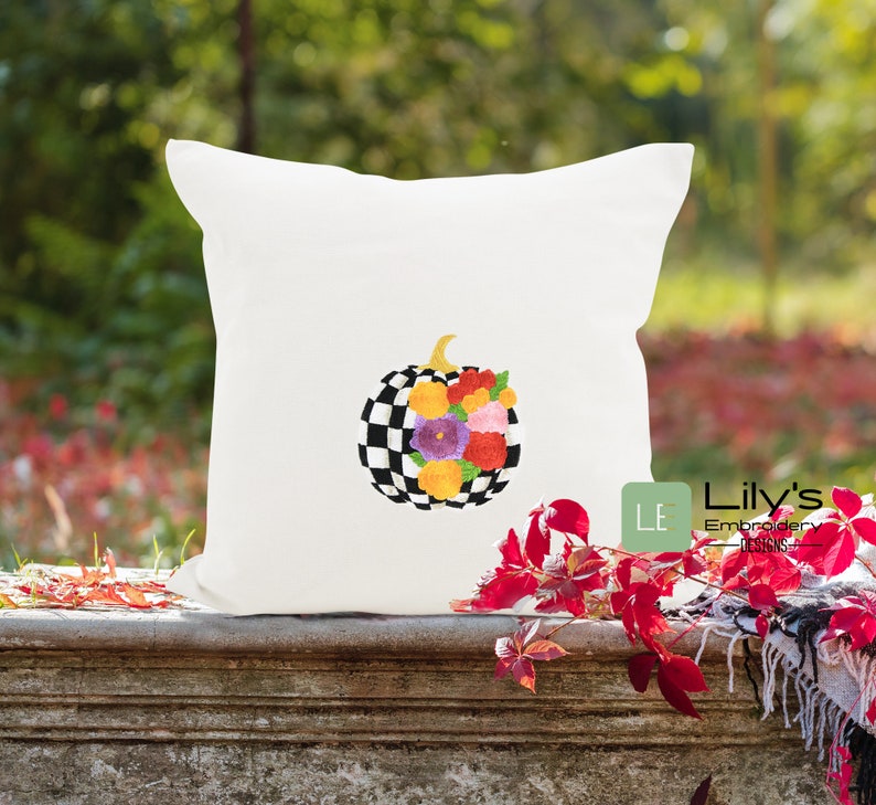 May include: A white cushion featuring a black and white checked pumpkin adorned with colourful flowers. The cushion is placed on a wooden surface with red leaves in the background. The text "Lily's Embroidery Designs" is visible in the bottom right corner of the image.