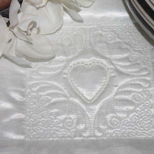 Peut inclure: Un tissu blanc carré et matelassé avec un motif en forme de cœur au centre. Le tissu a un aspect satiné et est orné de motifs floraux et tourbillonnants. Des fleurs d'orchidées blanches et un objet rayé noir et blanc sont en arrière-plan.