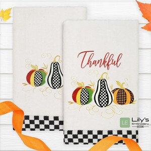 May include: Two white kitchen towels with black and white checkered borders. One towel features an embroidered design of three pumpkins with black and white checkered patterns and the word "Thankful" in red. The other towel features the same pumpkin design without the word "Thankful".