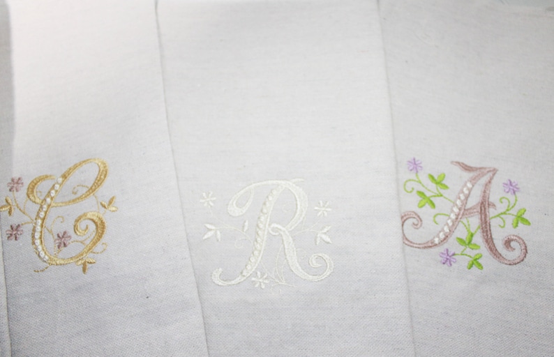 Puede incluir: Tres servilletas de lino blanco con iniciales bordadas. Las iniciales son "C" en dorado, "R" en blanco y "A" en malva, cada una con detalles florales. Las servilletas est&aacute;n dobladas y se muestran juntas.