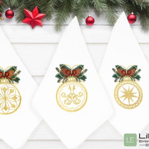 May include: Three white napkins with gold embroidered Christmas ornaments. Each ornament has a different design: a snowflake, a swirl, and a star. The ornaments are topped with a green and red bow.