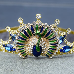 May include: A gold crown with a peacock design. The peacock is made of blue, green, and gold enamel and has clear rhinestones. The crown is decorated with clear rhinestones.