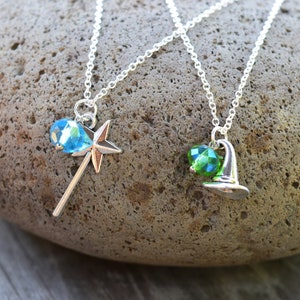 May include: Two silver necklaces with charm pendants. One features a star-shaped wand with a blue crystal, the other a witch hat with a green crystal. The necklaces are displayed on a textured stone surface.