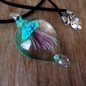 Pu&ograve; includere: Una collana con un ciondolo in vetro verde acqua con un fiore viola all'interno. Il ciondolo &egrave; su un cordoncino nero con una chiusura argentata e un ciondolo a forma di stella marina argentata.