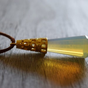 May include: A gold pendant with a clear, yellow-tinted crystal point hangs from a brown leather cord. The pendant has a detailed, ornate design.