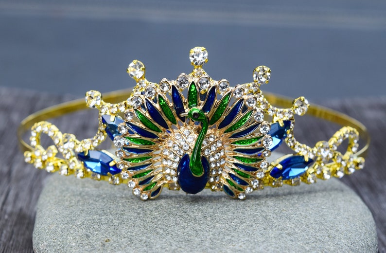 May include: A gold crown with a peacock design. The peacock is made of blue, green, and gold enamel and has clear rhinestones. The crown is decorated with clear rhinestones.