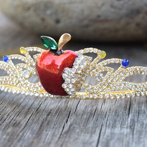 May include: Gold crown with a red apple and clear rhinestones. The apple has a green leaf and a bite taken out of it.