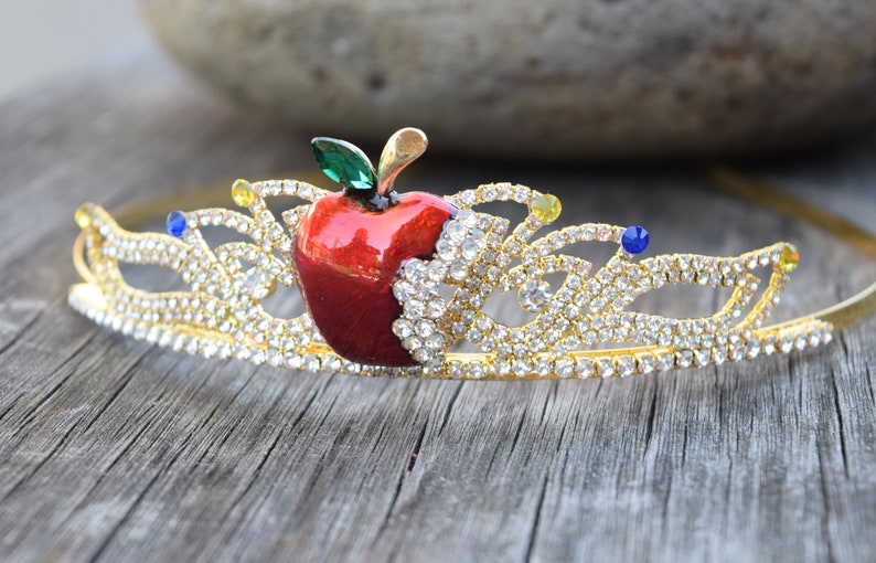 May include: Gold crown with a red apple and clear rhinestones. The apple has a green leaf and a bite taken out of it.