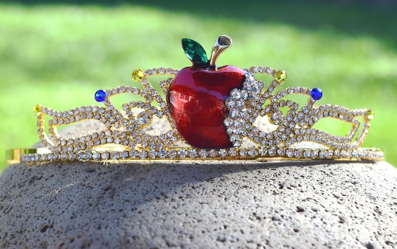 May include: A gold crown with a red apple in the center, surrounded by clear rhinestones. The apple has a green leaf and a gold stem.