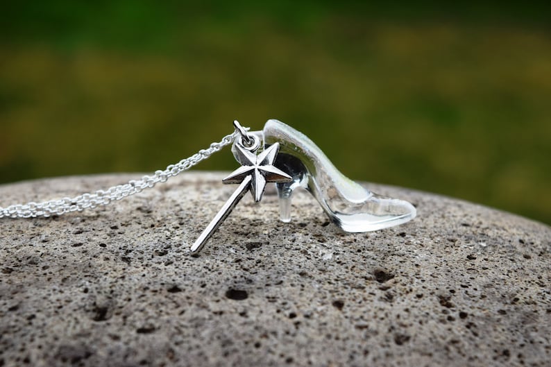 Pu&ograve; includere: Una collana d'argento con un ciondolo a forma di scarpetta di vetro e un ciondolo a forma di bacchetta magica con una stella in cima.