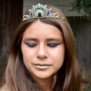 May include: A young person wearing a gold and blue peacock tiara with blue eyeliner. The tiara has a peacock design with blue and green feathers and rhinestones.