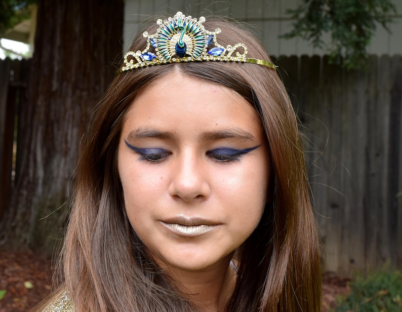 May include: A young person wearing a gold and blue peacock tiara with blue eyeliner. The tiara has a peacock design with blue and green feathers and rhinestones.