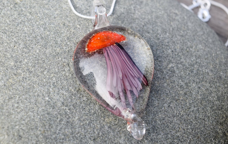 Pu&ograve; includere: Una collana con un ciondolo di vetro con un design a forma di medusa rossa e viola. Il ciondolo ha la forma di una lacrima e pende da una catena d'argento.