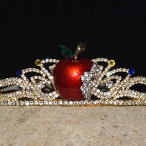 May include: A gold tiara with a red apple in the center. The tiara is decorated with clear rhinestones and has a few blue and yellow rhinestones.
