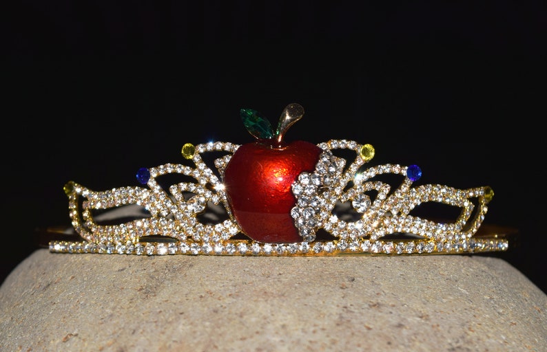 May include: A gold tiara with a red apple in the center. The tiara is decorated with clear rhinestones and has a few blue and yellow rhinestones.