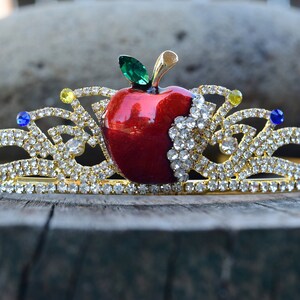 May include: A gold crown with a red apple in the center. The apple has a green leaf and is surrounded by clear rhinestones. The crown is decorated with blue and yellow rhinestones.