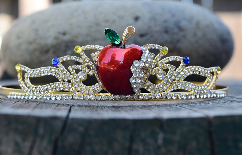 May include: A gold crown with a red apple in the center. The apple has a green leaf and is surrounded by clear rhinestones. The crown is decorated with blue and yellow rhinestones.