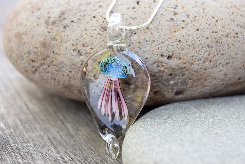 Pu&ograve; includere: Una collana con un ciondolo di vetro a forma di lacrima trasparente che contiene una medusa rosa e blu. Il ciondolo &egrave; appeso a una catena d'argento.