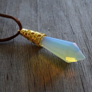 May include: A gold pendant with a clear iridescent gemstone in the shape of a teardrop. The pendant is attached to a brown leather cord.