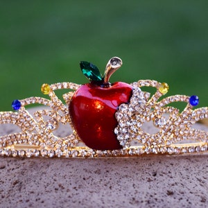 May include: A gold crown with a red apple and clear rhinestones. The apple has a green leaf and a bite taken out of it.