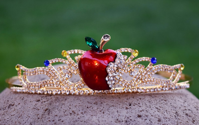 May include: A gold crown with a red apple and clear rhinestones. The apple has a green leaf and a bite taken out of it.