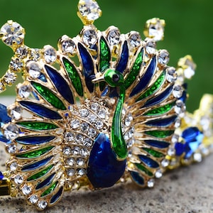 May include: A gold crown with a peacock design. The peacock is made of blue, green, and gold enamel and is surrounded by clear rhinestones.