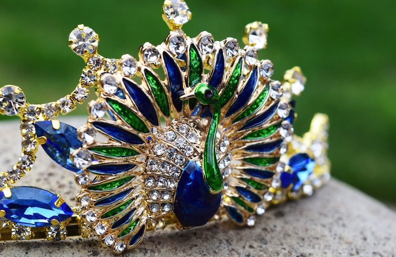 May include: A gold crown with a peacock design. The peacock is made of blue, green, and gold enamel and is surrounded by clear rhinestones.