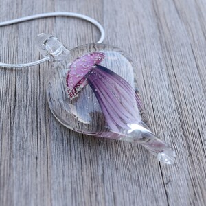 Pu&ograve; includere: Una collana con un ciondolo di vetro e una catena d'argento. Il ciondolo ha la forma di una lacrima e contiene un fiore viola con un centro rosa.