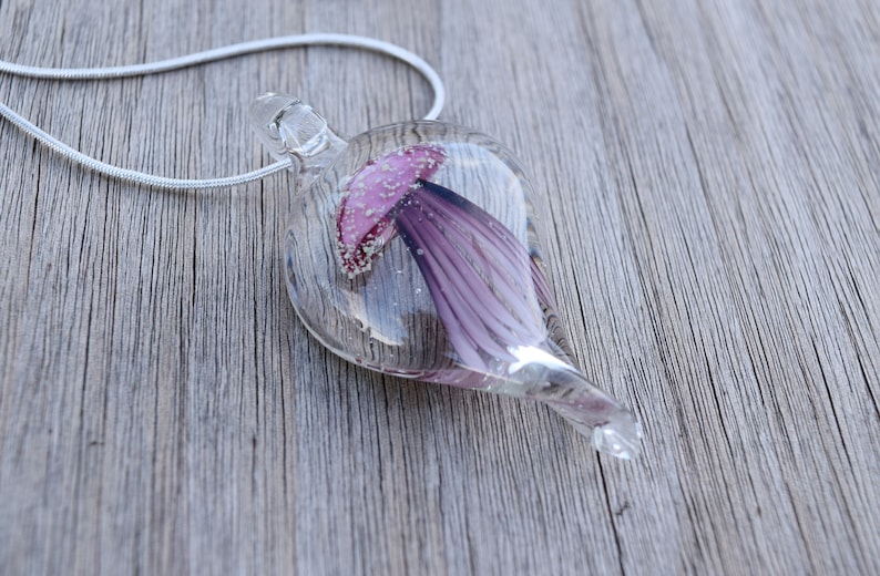 Pu&ograve; includere: Una collana con un ciondolo di vetro e una catena d'argento. Il ciondolo ha la forma di una lacrima e contiene un fiore viola con un centro rosa.