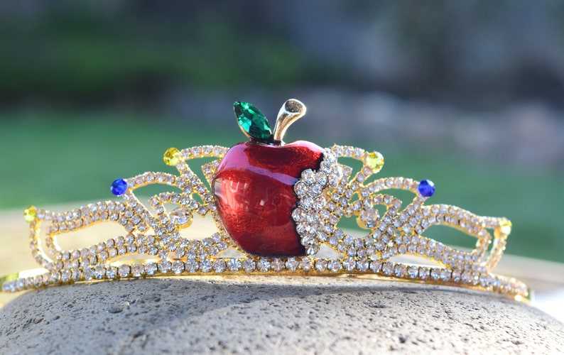 May include: A gold crown with a red apple and green leaf design. The crown is decorated with clear rhinestones.