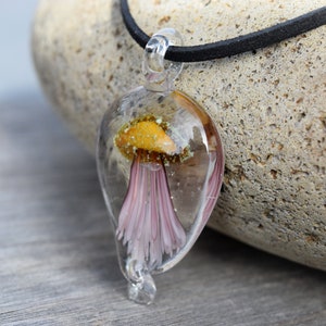 Pu&ograve; includere: Una collana con un ciondolo in vetro trasparente che contiene un delicato fiore rosa incapsulato in resina. Il ciondolo ha la forma di una lacrima e pende da un cordoncino in pelle nera.