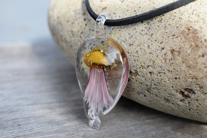 Pu&ograve; includere: Una collana con un ciondolo in vetro trasparente che contiene un delicato fiore rosa incapsulato in resina. Il ciondolo ha la forma di una lacrima e pende da un cordoncino in pelle nera.