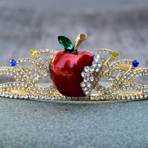 May include: A gold crown with a red apple and clear rhinestones. The apple has a green leaf and a bite taken out of it.