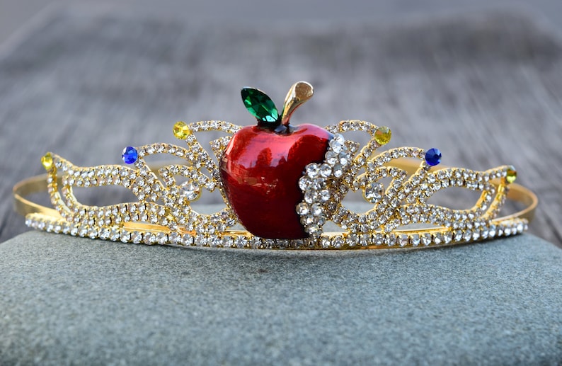 May include: A gold crown with a red apple and clear rhinestones. The apple has a green leaf and a bite taken out of it.