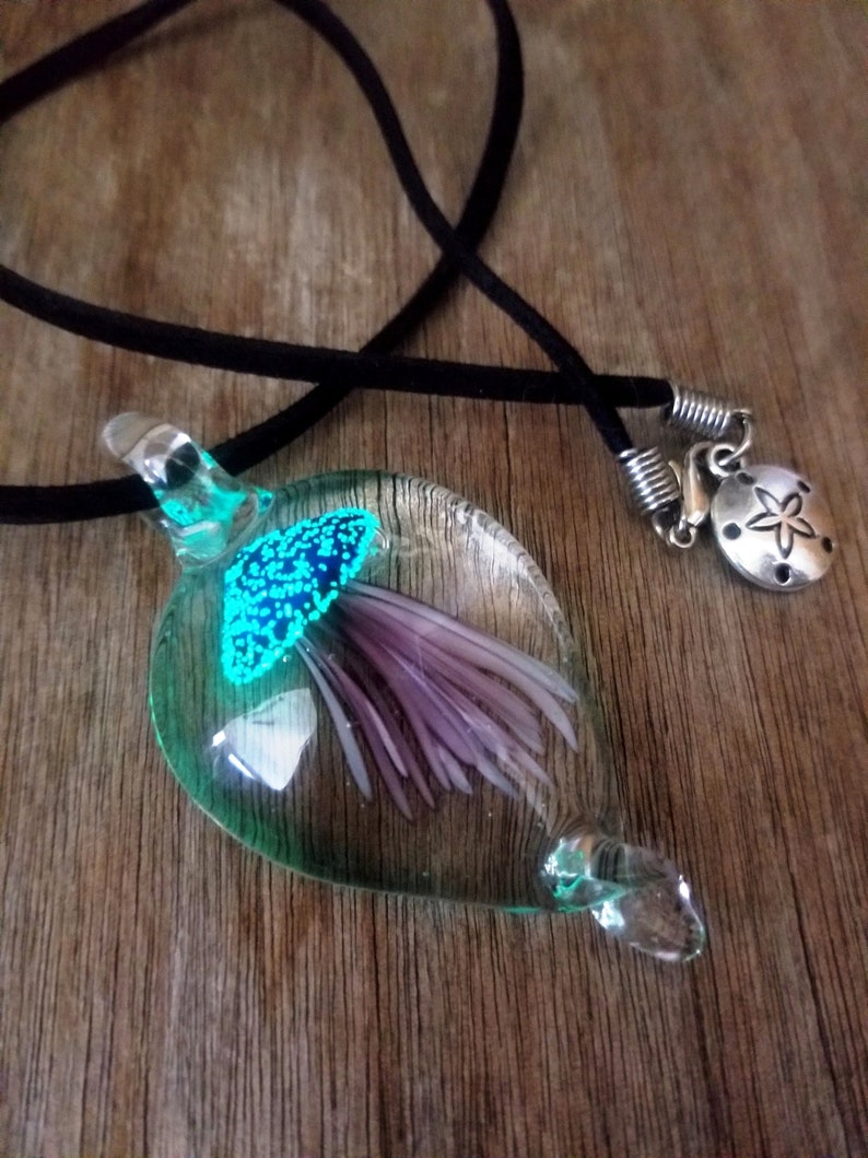 Pu&ograve; includere: Una collana con un ciondolo in vetro verde acqua con un fiore viola all'interno. Il ciondolo &egrave; su un cordoncino nero con una chiusura argentata e un ciondolo a forma di stella marina argentata.