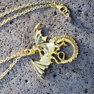 May include: A gold chain necklace with a dragon pendant. The dragon is curled up with its wings spread out. The necklace has a small purple gemstone on the clasp.