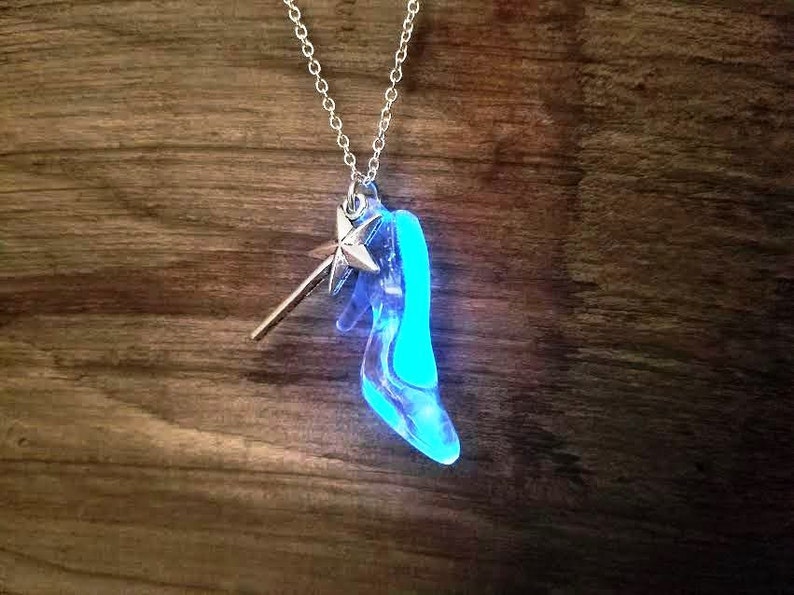 Pu&ograve; includere: Una collana d'argento con un ciondolo a forma di scarpetta di vetro blu e un ciondolo a forma di bacchetta magica d'argento. Il ciondolo a forma di scarpetta brilla di blu.