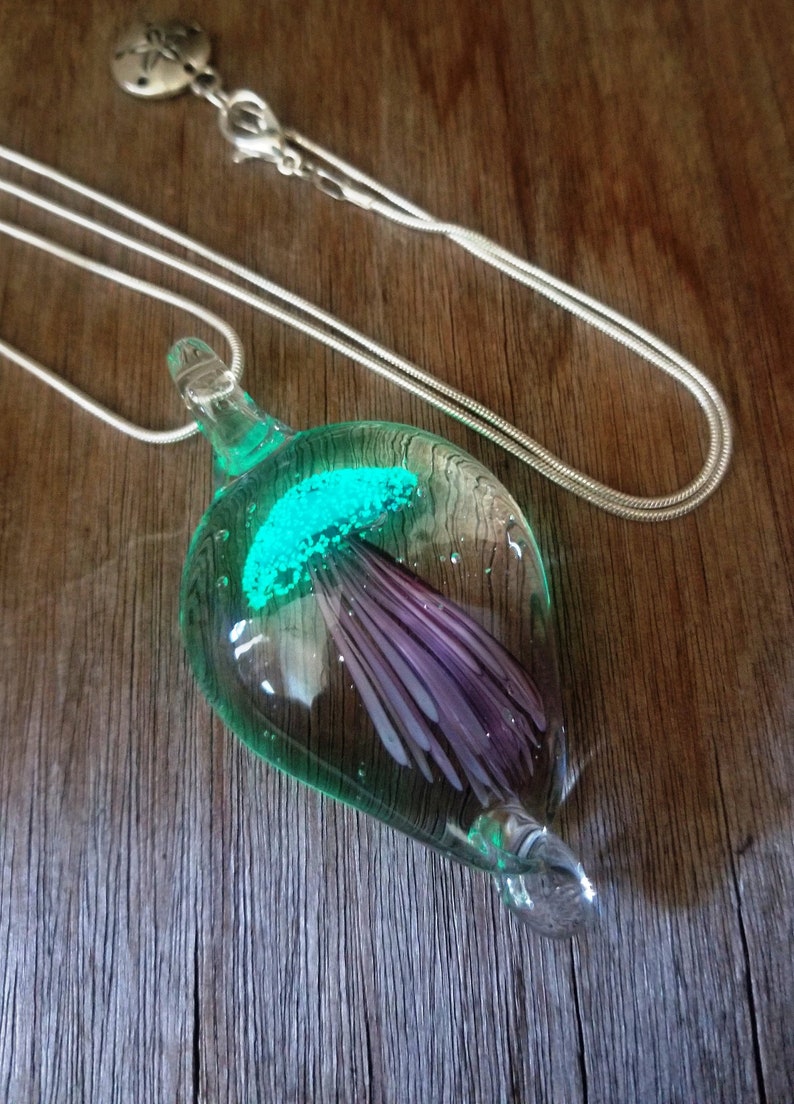 Pu&ograve; includere: Una collana con catena d'argento con un ciondolo di vetro che presenta una medusa viola e verde al suo interno. La medusa brilla di verde.
