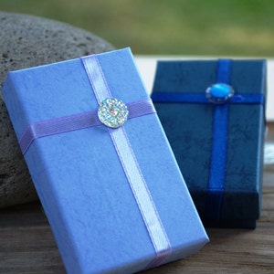 May include: Two small gift boxes with ribbon and decorative embellishments. The box on the left is light blue with a lavender ribbon and a clear rhinestone. The box on the right is dark blue with a navy blue ribbon and a blue rhinestone.