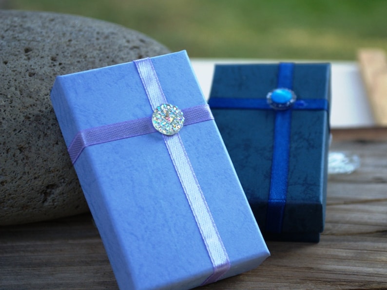 May include: Two small gift boxes with ribbon and decorative embellishments. The box on the left is light blue with a lavender ribbon and a clear rhinestone. The box on the right is dark blue with a navy blue ribbon and a blue rhinestone.
