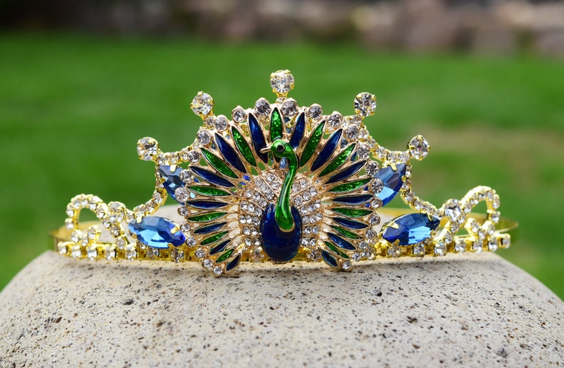May include: A gold crown with a peacock design. The peacock is made of blue, green, and gold enamel. The crown is decorated with clear and blue rhinestones.