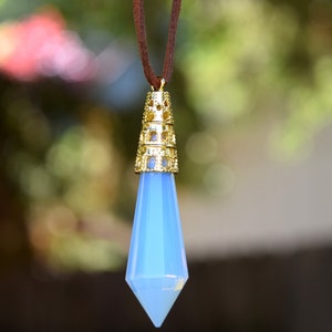 May include: A light blue crystal pendant with a gold filigree setting hangs from a brown cord. The pendant is shaped like a teardrop and has a pointed tip.