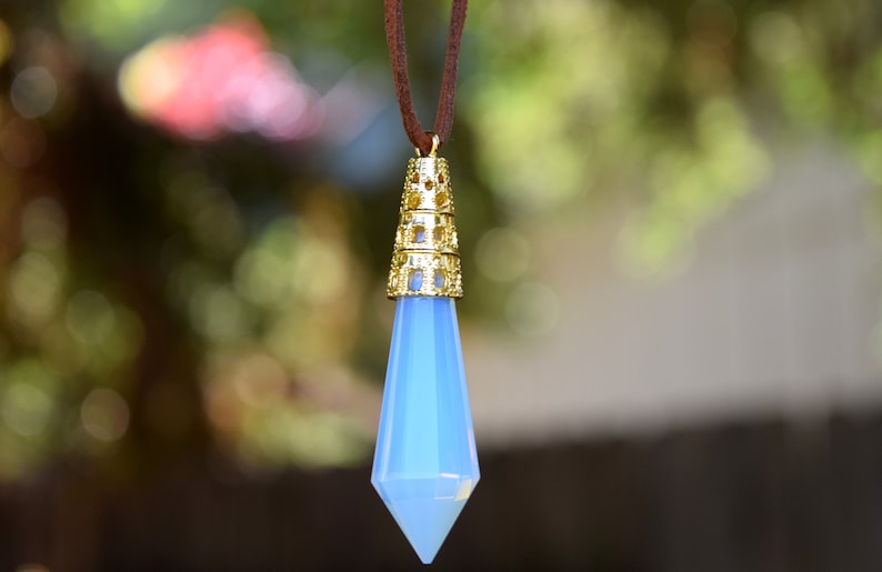 May include: A light blue crystal pendant with a gold filigree setting hangs from a brown cord. The pendant is shaped like a teardrop and has a pointed tip.