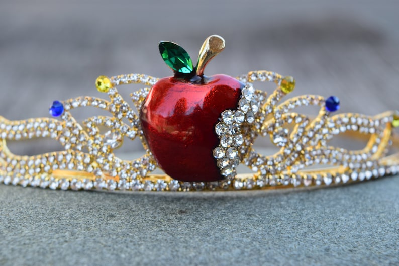 May include: A gold crown with a red apple and clear rhinestones. The apple has a green leaf and a bite taken out of it.