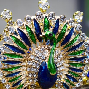 May include: A gold crown with a peacock design. The peacock is made of blue, green, and gold enamel. The crown is decorated with clear rhinestones.