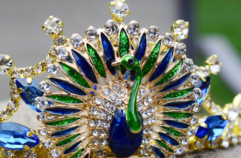 May include: A gold crown with a peacock design. The peacock is made of blue, green, and gold enamel. The crown is decorated with clear rhinestones.