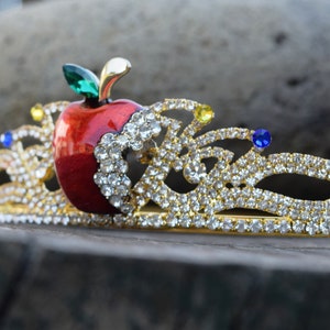 May include: A gold-tone tiara with a red apple and clear rhinestones. The apple has a green leaf and is surrounded by a gold-tone crown design.