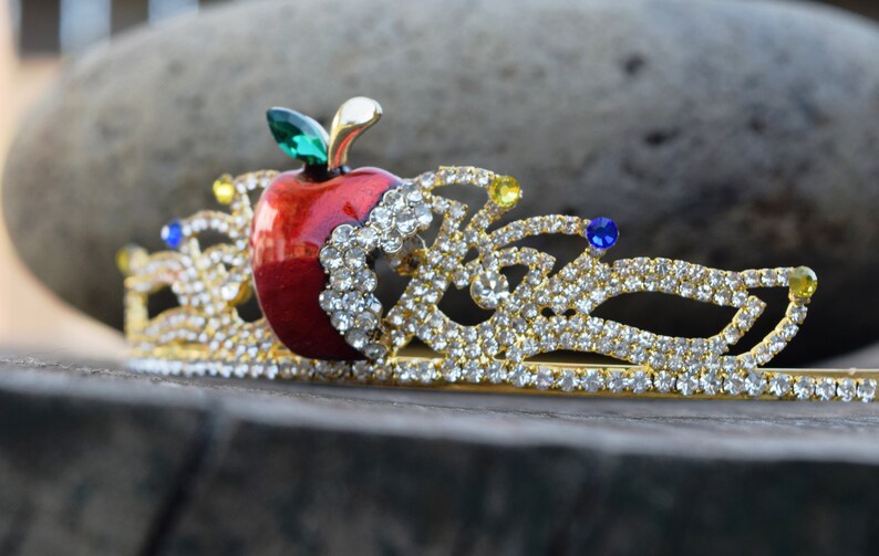 May include: A gold-tone tiara with a red apple and clear rhinestones. The apple has a green leaf and is surrounded by a gold-tone crown design.