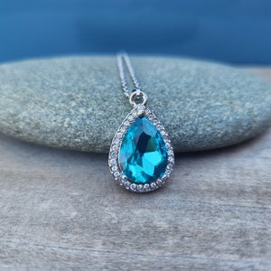 Puede incluir: Un collar con un colgante en forma de l&aacute;grima adornado con una gran piedra preciosa azul brillante rodeada por un halo de peque&ntilde;as piedras preciosas transparentes. El colgante es plateado y cuelga de una delicada cadena de plata.
